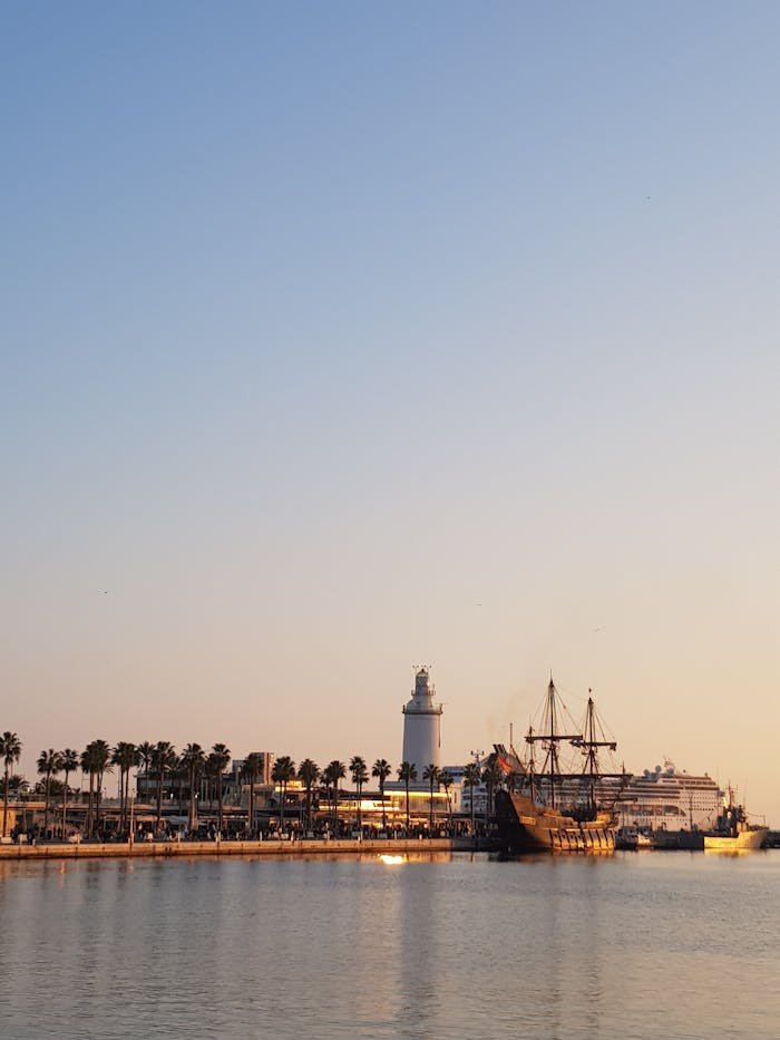 Scenic view of Málaga port at sunset featuring La Farola lighthouse and historic ships.